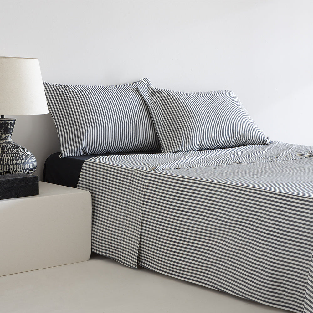 Juego de sábanas de algodón a rayas azules compuesto por sábana encimera, bajera y fundas de almohada a juego, presentado sobre una cama de matrimonio en un dormitorio luminoso y moderno, destacando la suavidad, transpirabilidad, resistencia y diseño clásico del tejido.